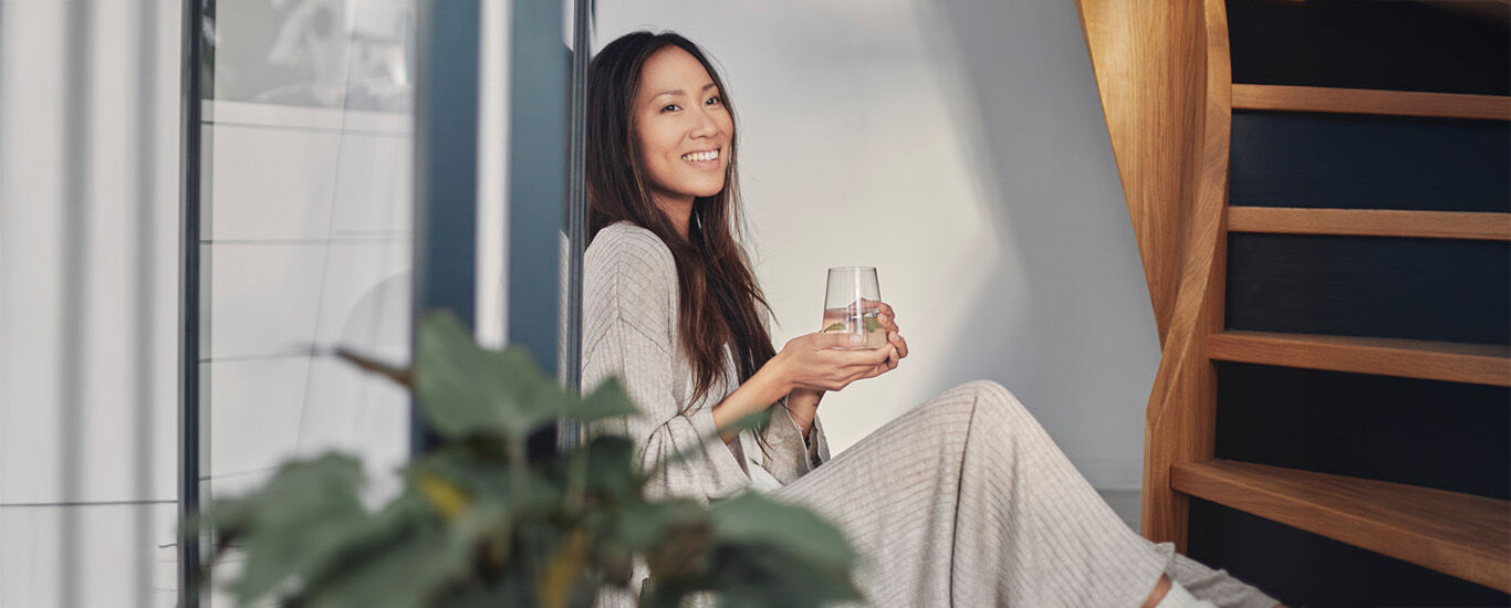 Frau sitzt angelehnt an einem Fenster mit einem Glas Wasser in der Hand.