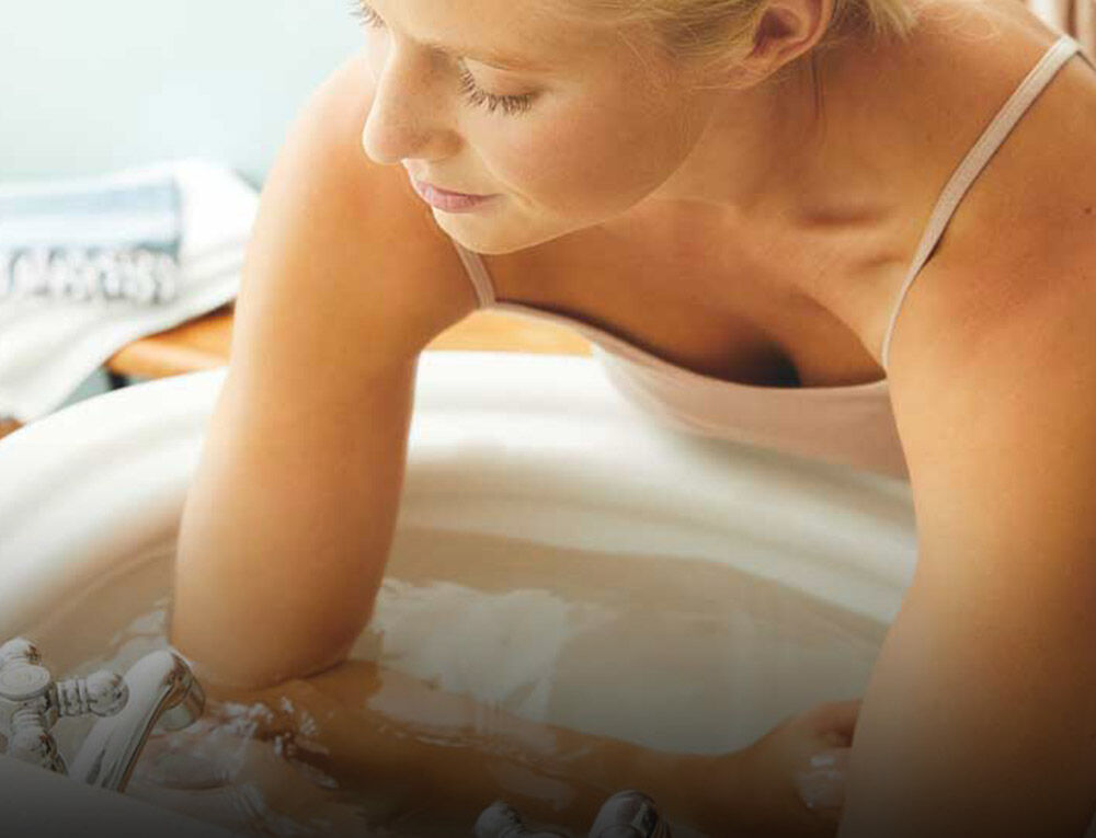 Blonde Frau in Top führt ein kaltes Armbad im Waschbecken durch. 
