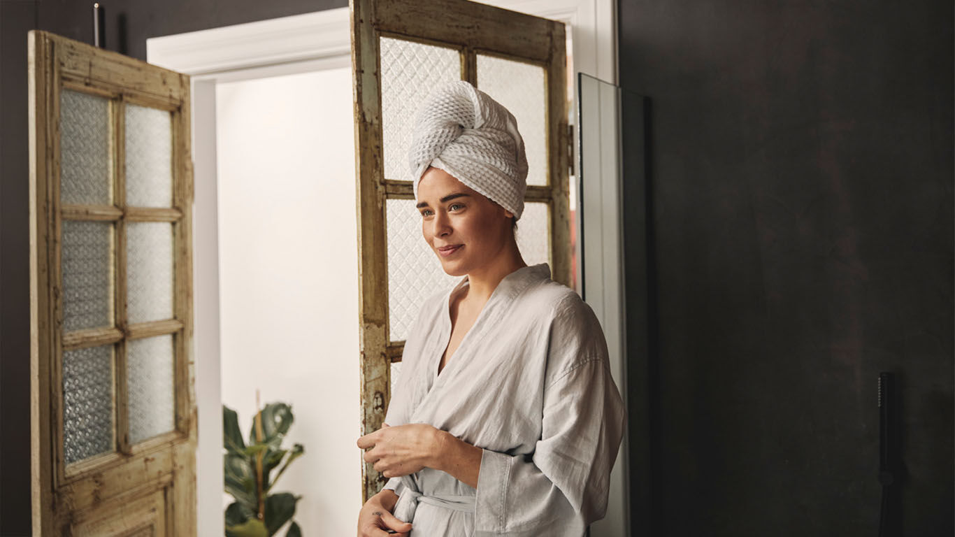 Frau in einem hellen Badezimmer tr&auml;gt einen Bademantel und ein Handtuch um den Kopf, steht vor einer ge&ouml;ffneten Holzt&uuml;r.