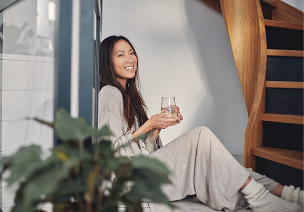 Frau in Loungewear sitzt auf dem Boden und lehnt an der Wand. In der Hand h&auml;lt sie ein Glas.
