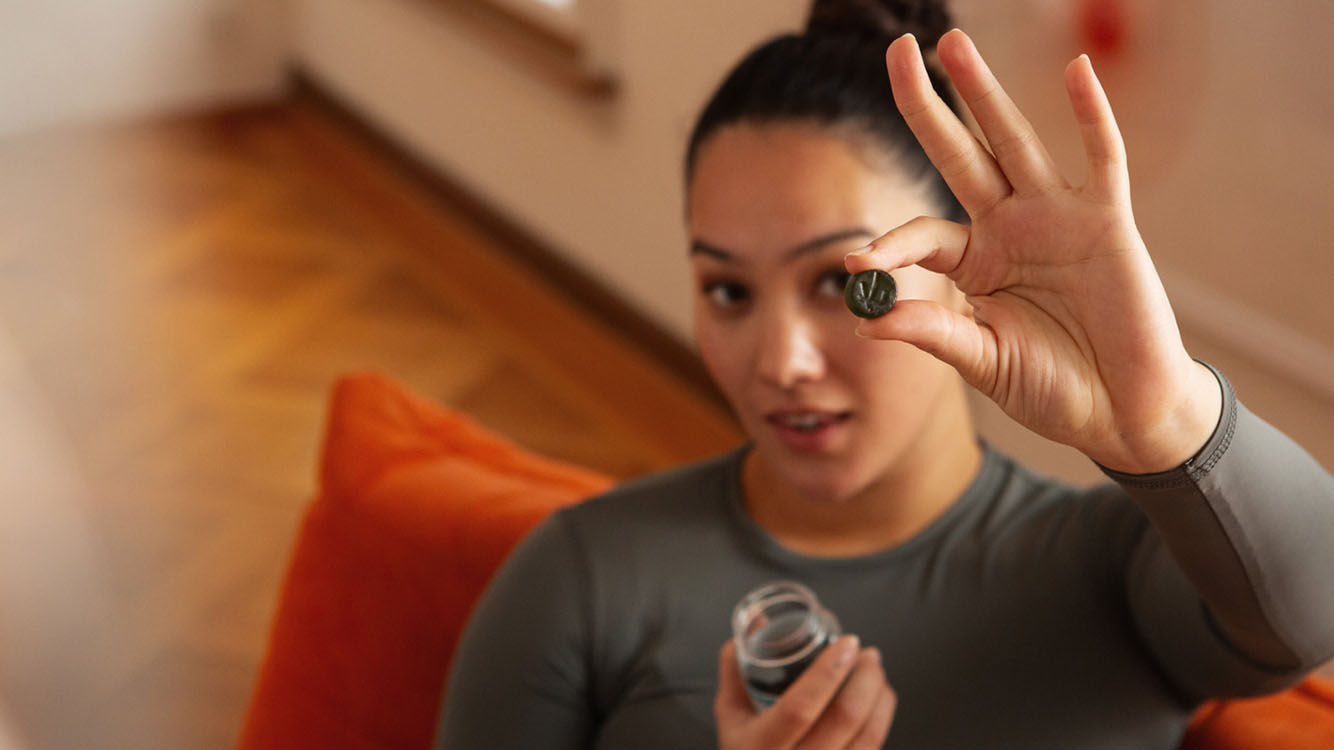 Frau hebt eine Hand mit einem Kneipp Gummie in die Kamera und h&auml;lt in der anderen eine Kneipp Gummie-Verpackung, sitzend auf einem orangefarbenen Sofa.