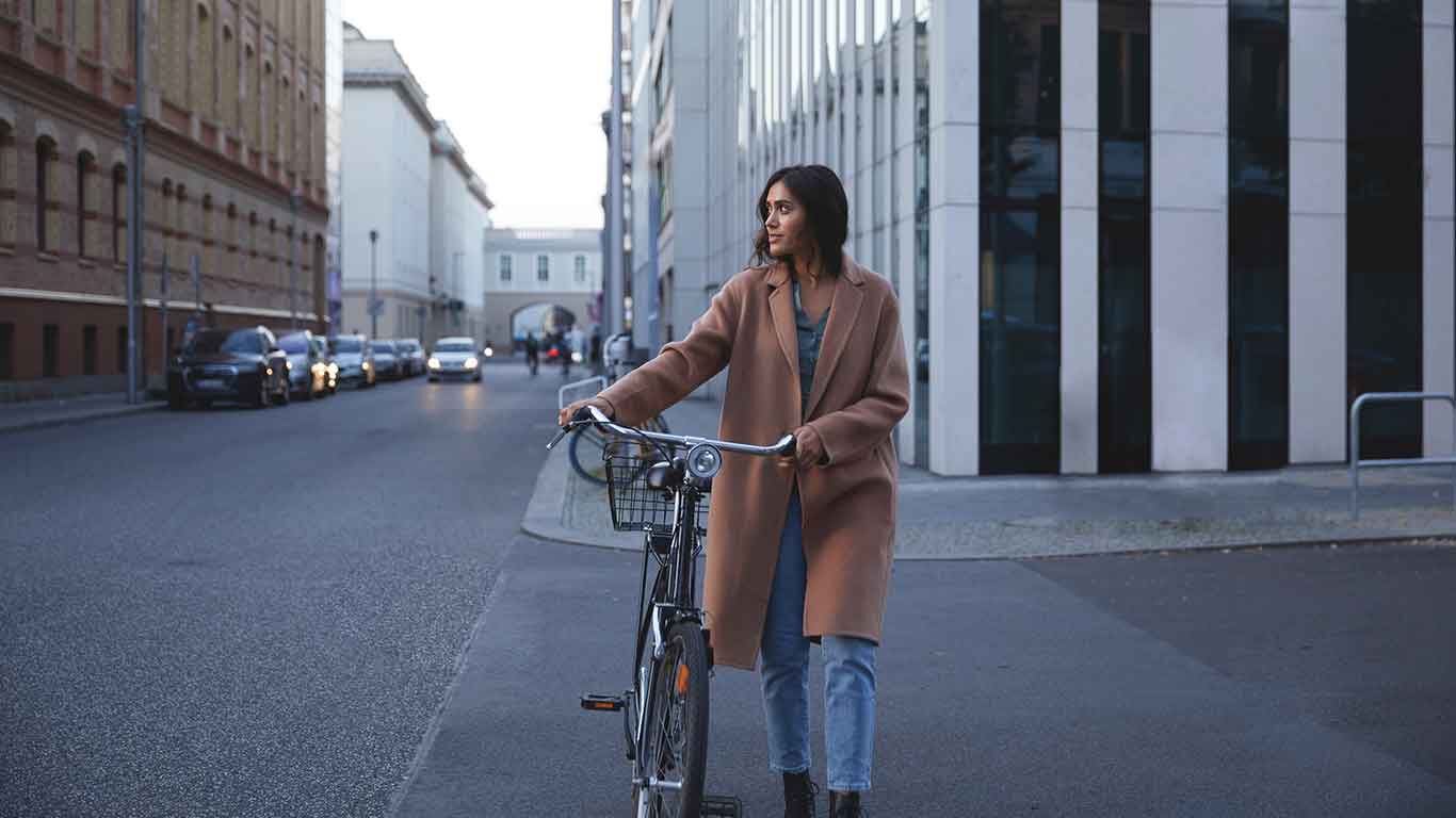 Frau in braunem Wollmantel schiebt Fahrrad über die Straße.