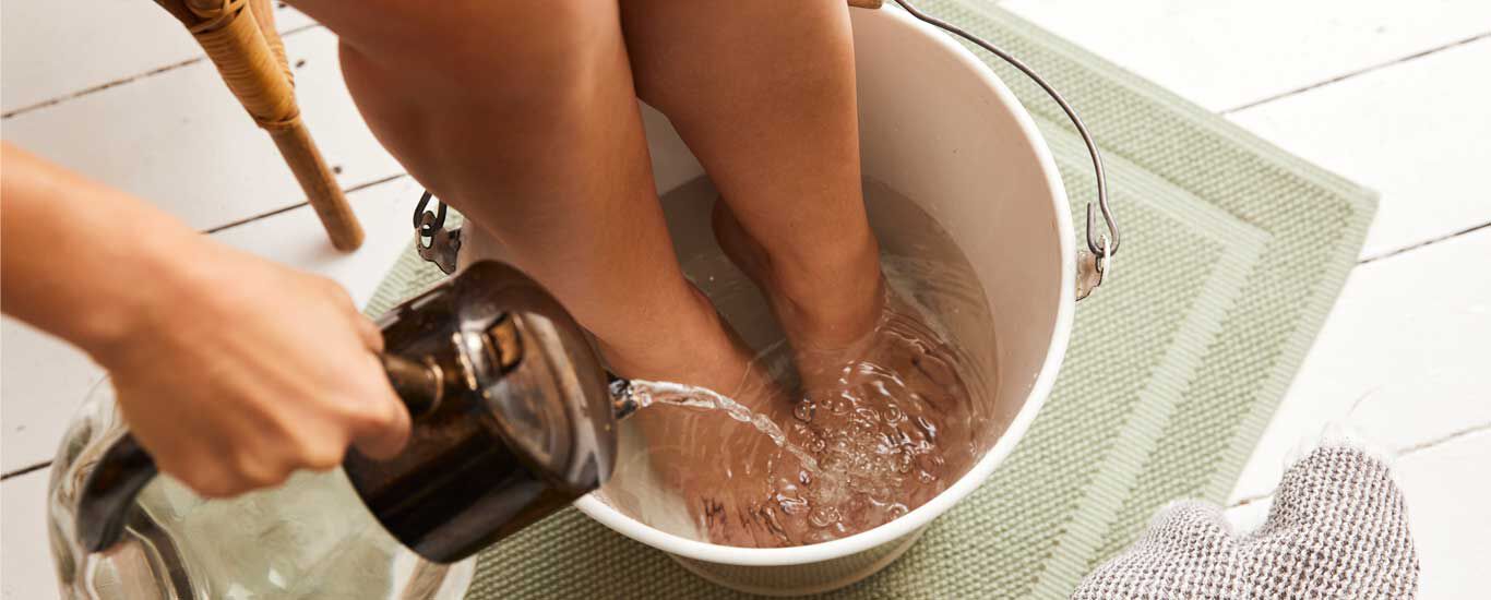 Eine Frau hat ihre Füße in einem mit Wasser gefüllten Eimer und gießt weiteres Wasser mit einer Kanne in den Eimer.