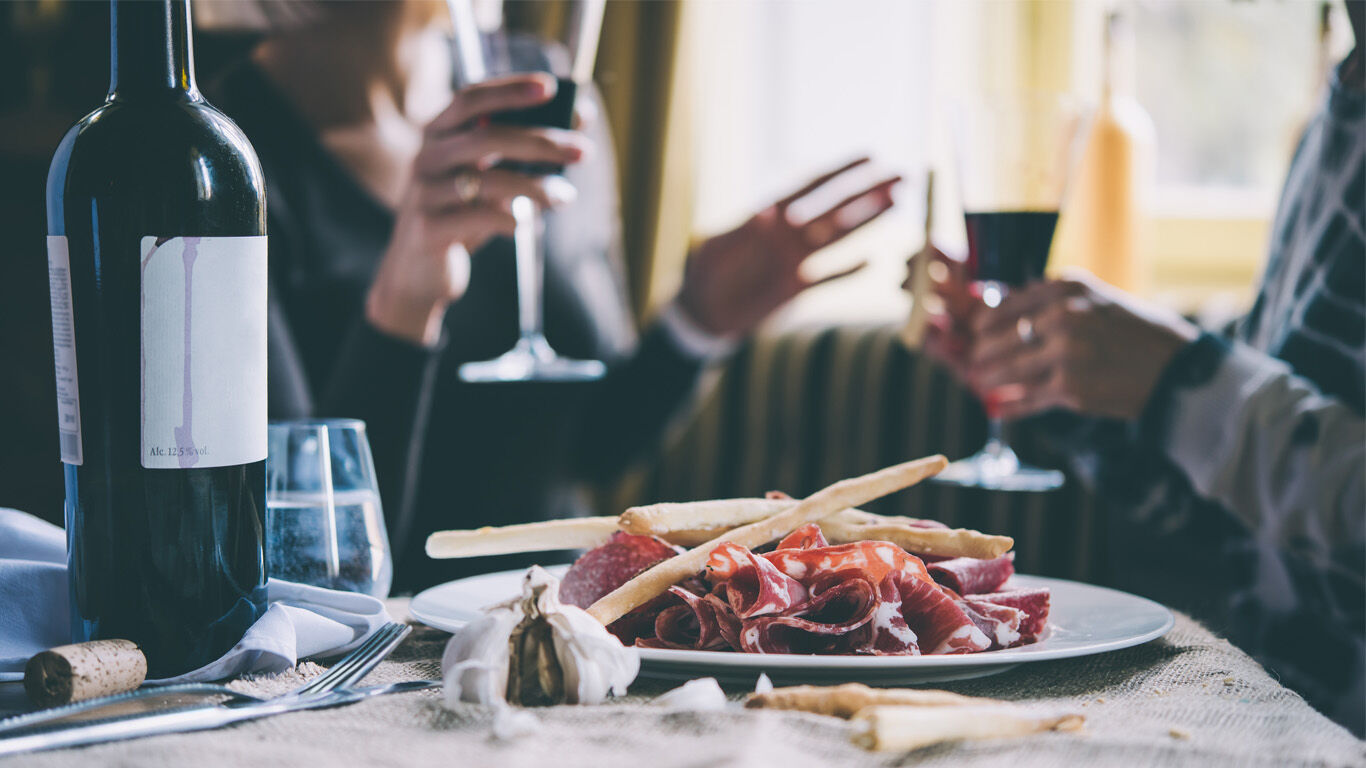 Antipasti Platte neben einer Weinflasche, im Hintergrund 2 Personen mit Weingläsern in der Hand.