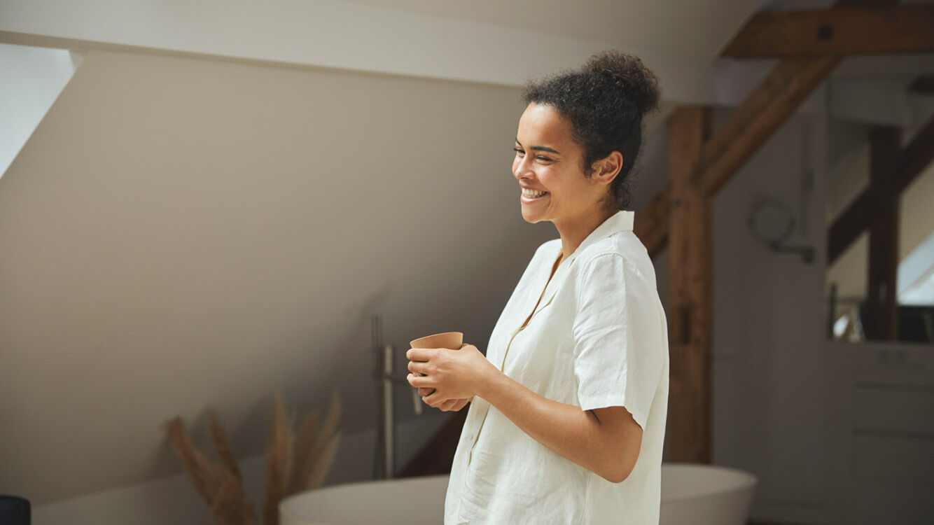 Frau steht entspannt in einem hellen, modernen Raum und h&auml;lt eine Tasse in der Hand.