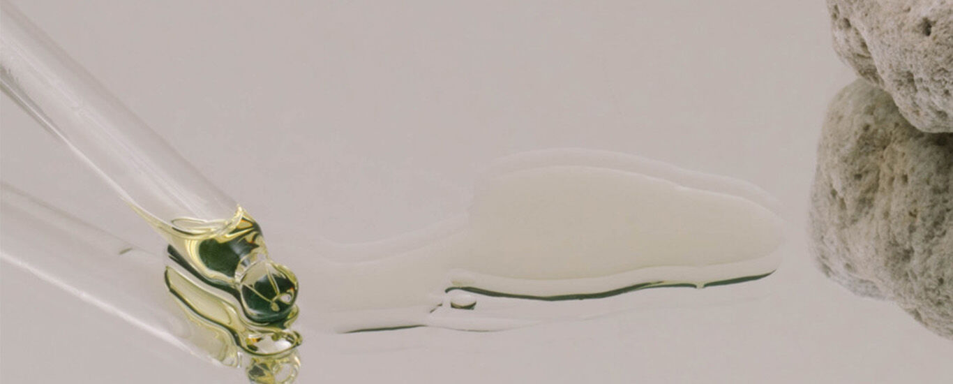 Pipette mit einem Tropfen klarer Flüssigkeit neben einem porösen Stein auf spiegelnder Oberfläche.