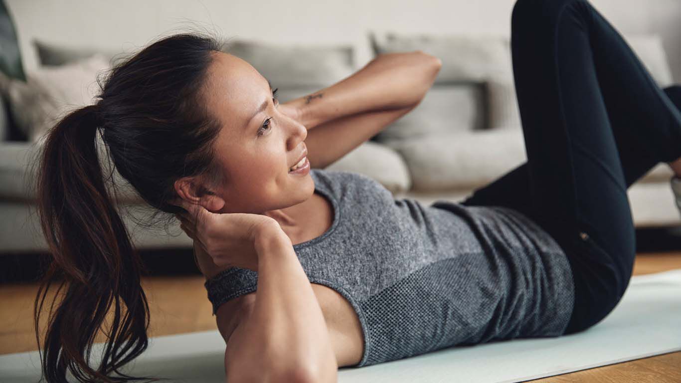Frau liegt auf einer Sportmatte im Wohnzimmer und macht Sit-ups.