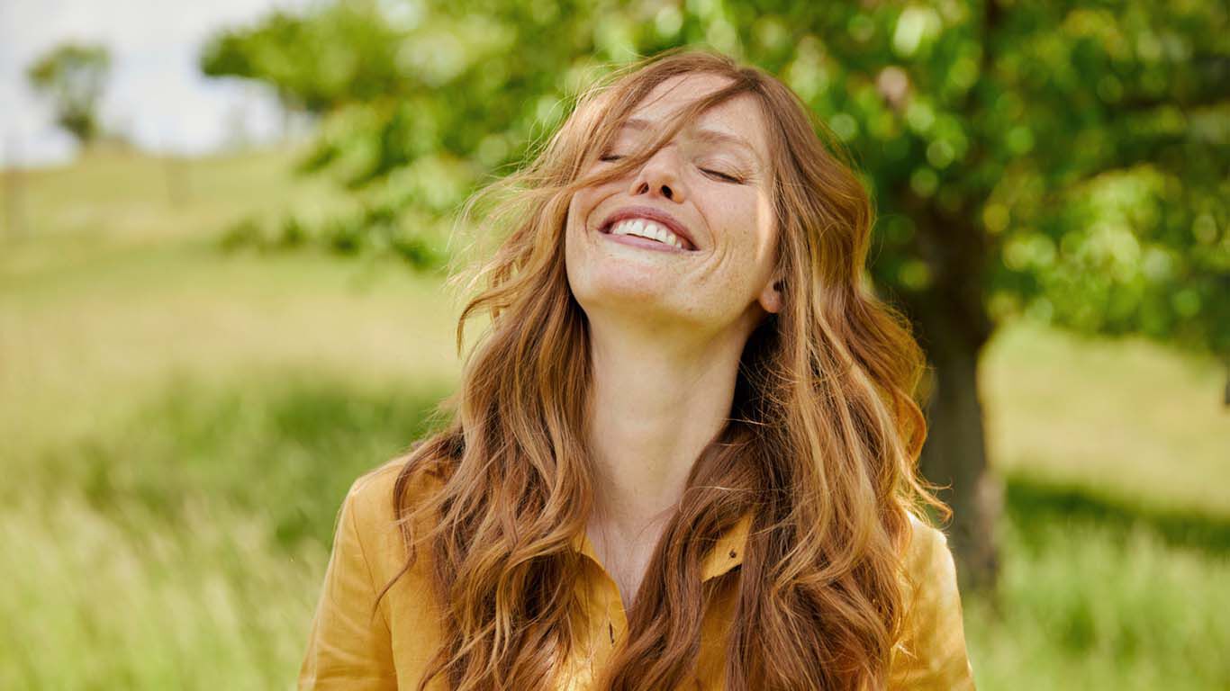 Frau stehl herzlich lachend auf einer Wiese und hat ihre haare ins Gesicht hängen.