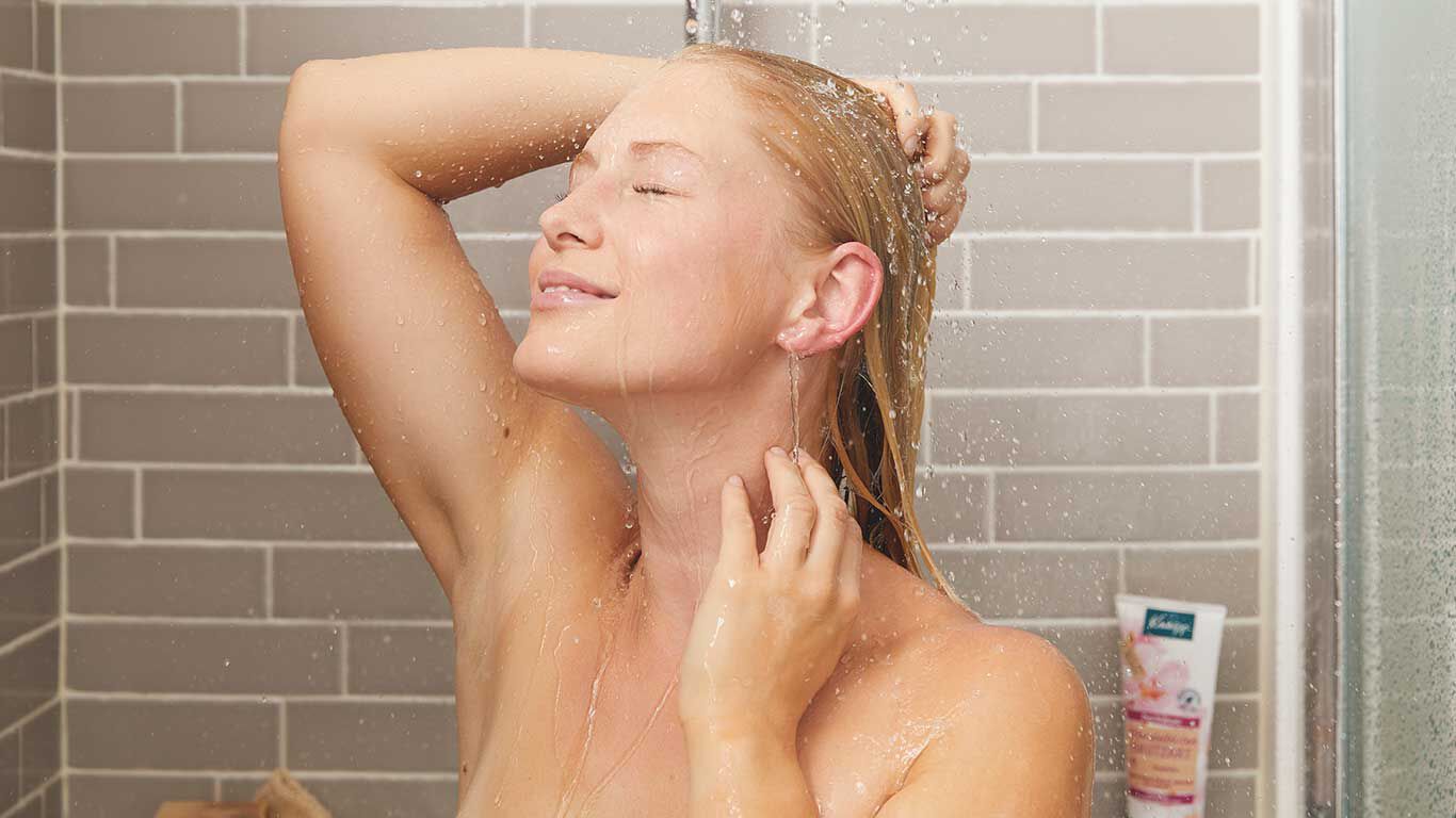 Frau steht unter der Dusche hält ihr Gesicht unter den Duschstrahl.