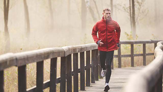 Person in roter Laufjacke joggt auf einem Holzsteg durch einen nebligen Wald.