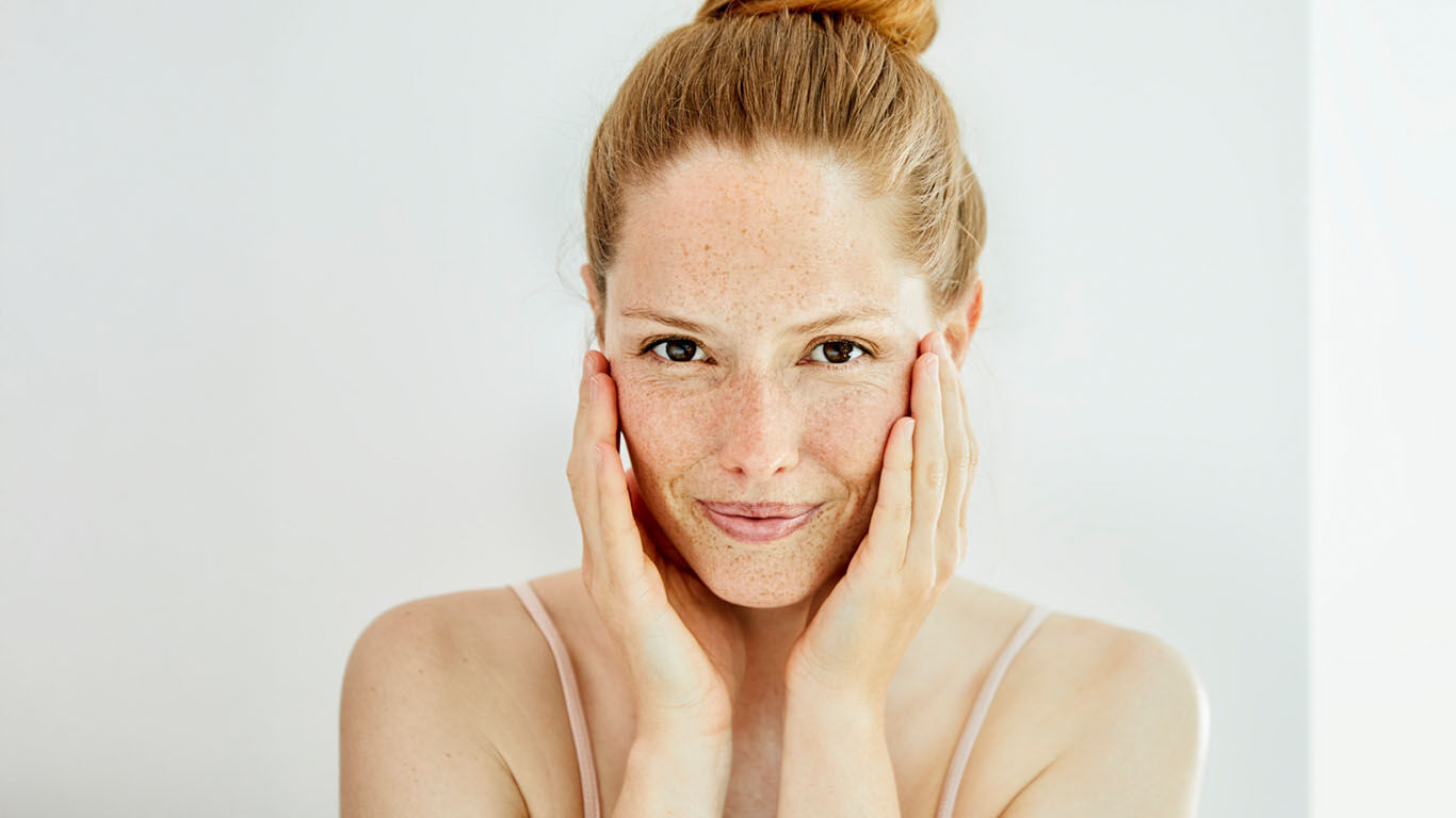 Frau steht mit zusammengebundenen Haaren vor einem Siegel und hat die Hände am GEsicht.