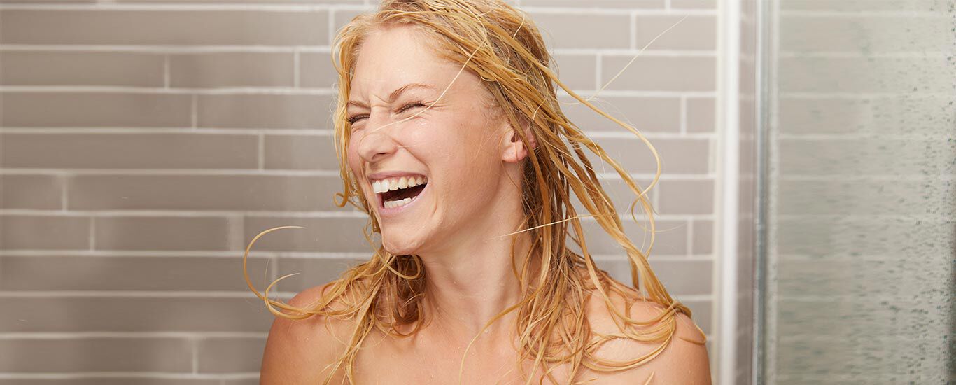Blonde Frau in der Dusche. Ihre nassen Haare wirft sie lachend um sich.