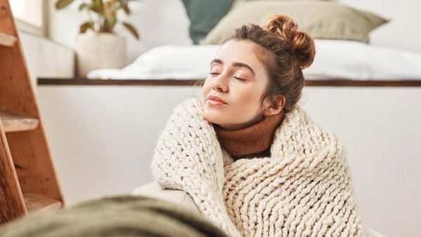 Frau mit Dutt sitzt in einer Kuscheldecke eingewickelt auf dem Boden und genießt die Ruhe.