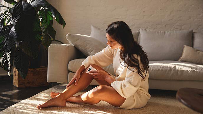 Une femme aux cheveux noirs est assise sur un tapis et applique de la crème sur ses jambes. 