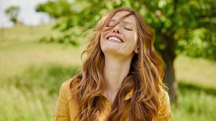 Une femme rousse en chemisier jaune se tient dans une prairie verte, renverse sa tête en arrière et rit.