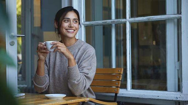 Frau sitzt mit einer Tasse Heißgetränk auf einem Holzstuhl bei geöffnetem Fenster.