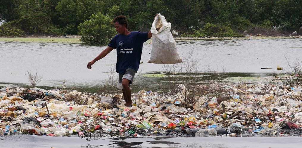 ReSea Angestellter steht in einem Berg Plastikmüll und hält einen vollen Sack mit Plastikmüll in die Höhe.