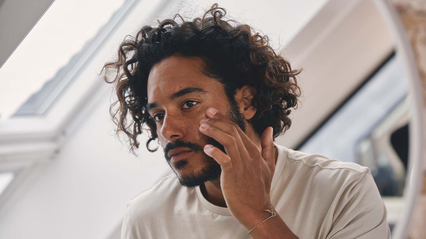 Un homme se tient devant le miroir dans la salle de bain et applique de la crème sur son visage. 