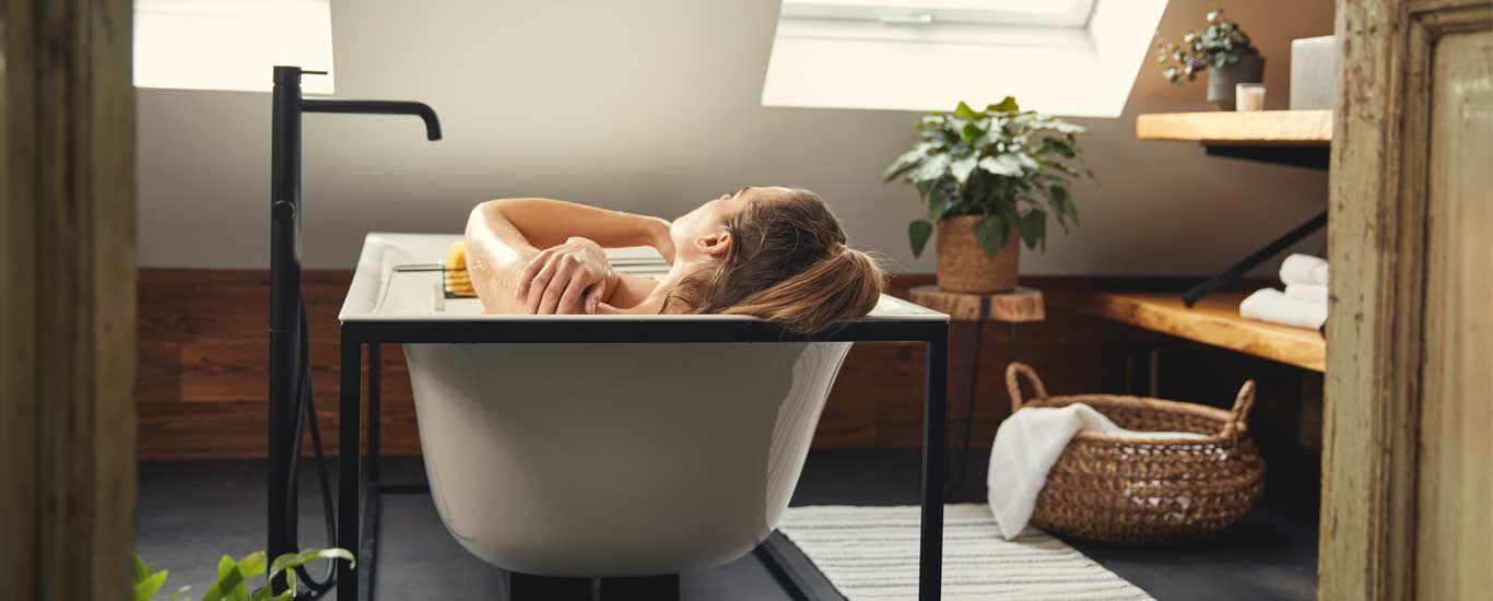 In einem Badezimmer liegt eine Frau in einer Badewanne, man kann sie nur von hinten sehen.