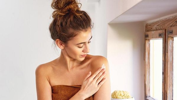 Une femme aux cheveux foncés, enveloppée dans une serviette, applique de la crème sur son épaule. 