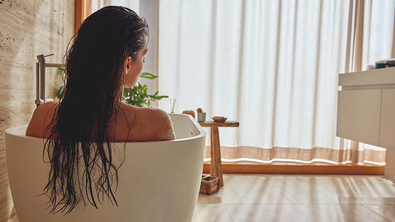 Frau mit langen braunen Haaren entspannt in einer Badewanne. Ansicht von hinten.
