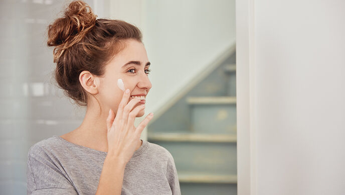 Une femme aux cheveux bruns et au chignon applique de la crème sur son visage en souriant.