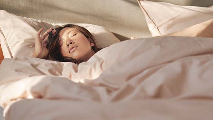 Dark haired woman in white t-shirt sleeps in bed with beige linen cover.