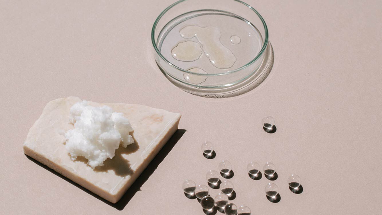 Boîte de Pétri remplie de liquide, avec à côté des billes transparentes et du beurre de karité sur un morceau de marbre. 