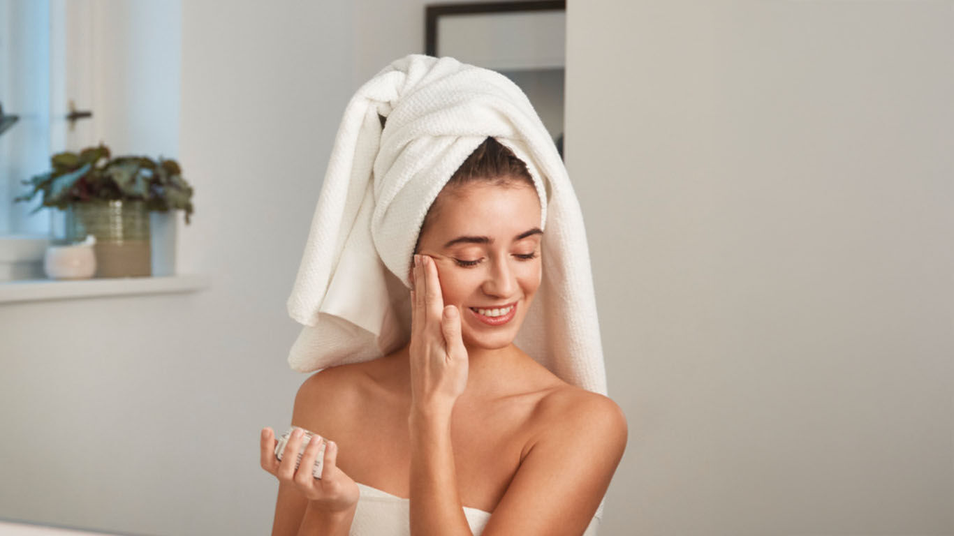 Une femme se tient devant le miroir de la salle de bain avec une serviette sur la tête et s'applique de la crème sur le visage.