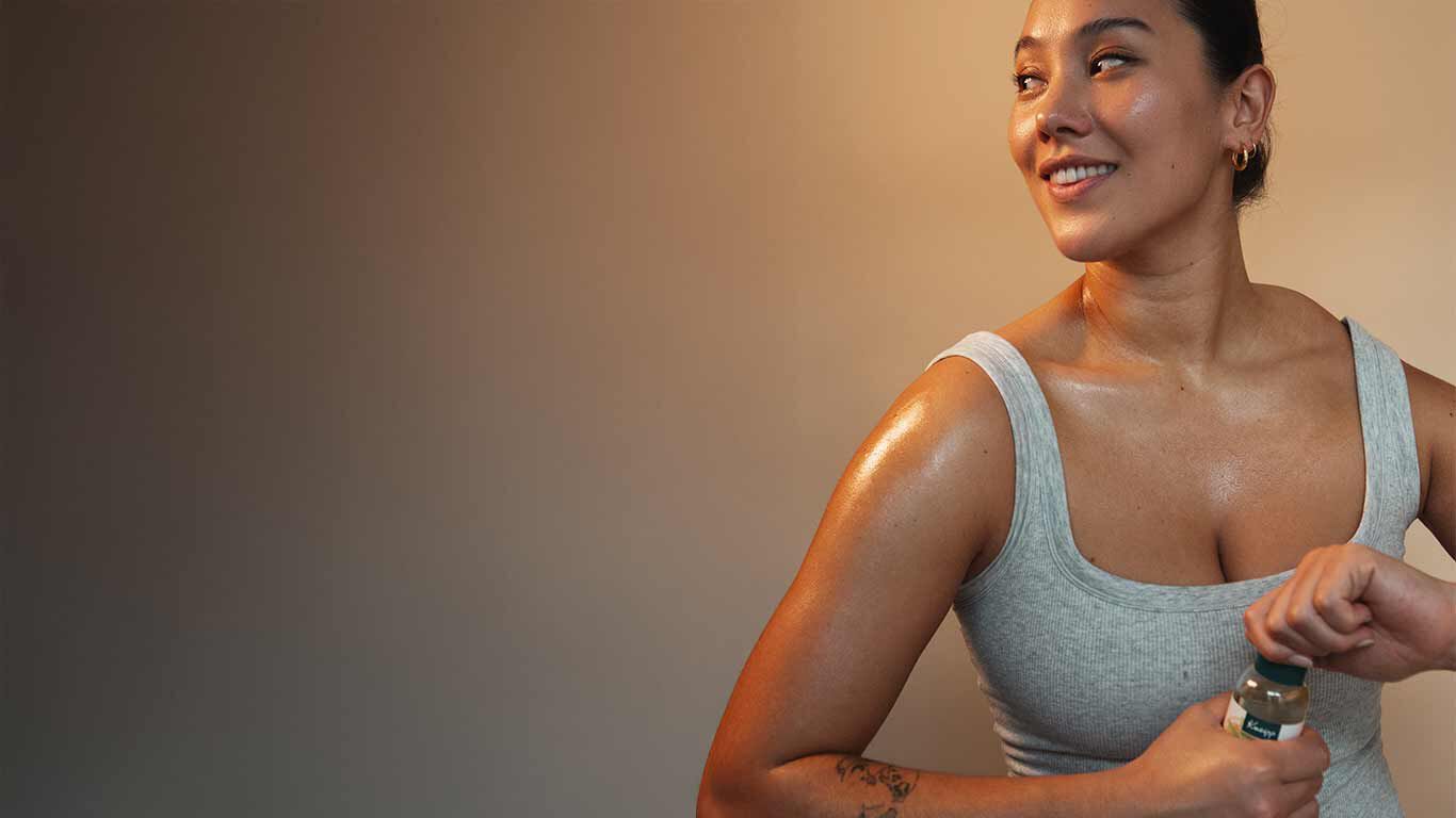 Une femme en haut gris tient une bouteille d'huile Kneipp dans sa main. Elle dévisse le flacon. 