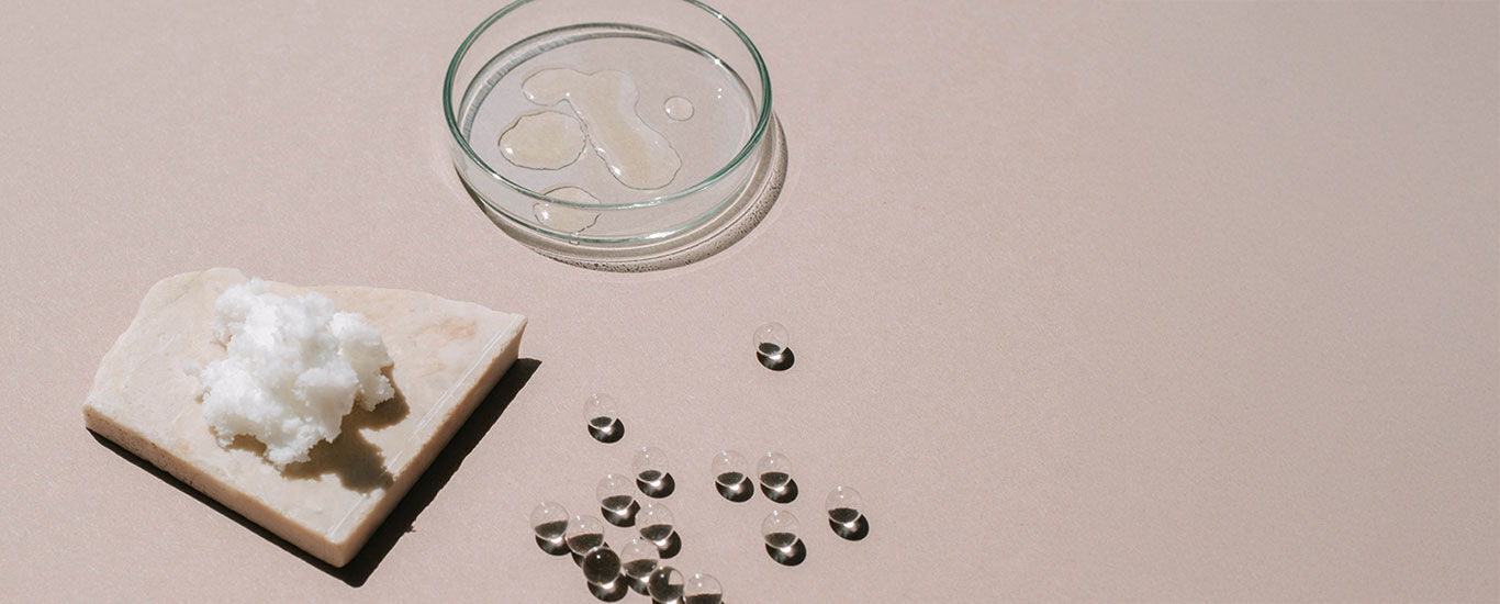 Bo&icirc;te de P&eacute;tri contenant un liquide transparent, &agrave; c&ocirc;t&eacute; du beurre de karit&eacute; sur un morceau de pierre et des perles de verre &agrave; c&ocirc;t&eacute;.