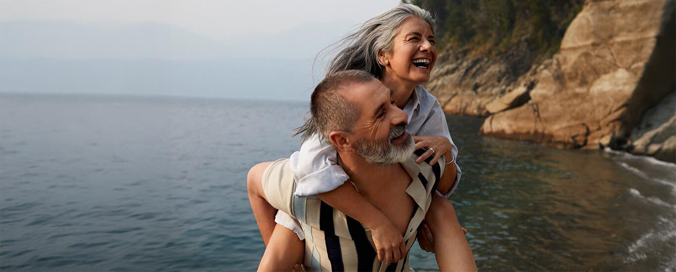 Un homme âgé porte une femme âgée sur son dos sur la plage. En arrière-plan, des parois rocheuses. Tous deux rayonnent de joie.