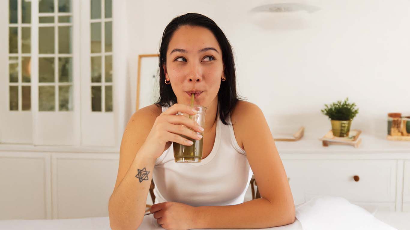 Femme en haut blanc avec de longs cheveux noirs, appuyée sur le comptoir de la cuisine, buvant un smoothie vert dans un verre avec une paille. 