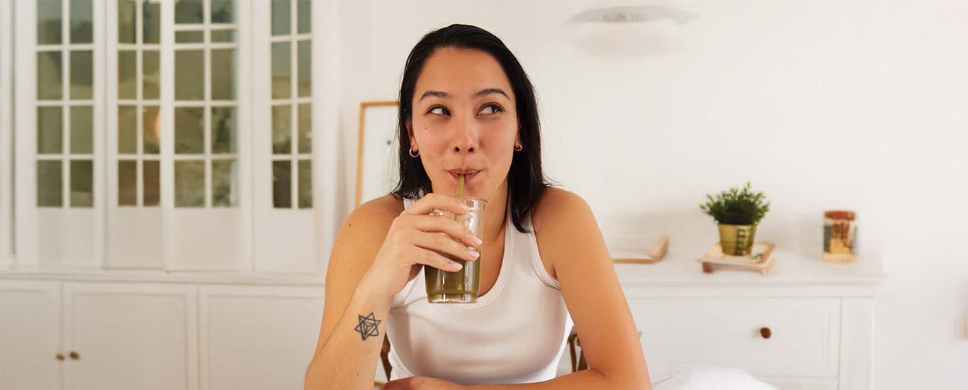 Femme en haut blanc avec de longs cheveux noirs, appuyée sur le comptoir de la cuisine, buvant un smoothie vert dans un verre avec une paille. 