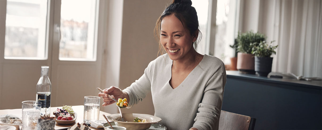 Une femme avec un chignon est assise &agrave; la table du repas, elle tient une cuill&egrave;re &agrave; la main et rit de bon c&oelig;ur.