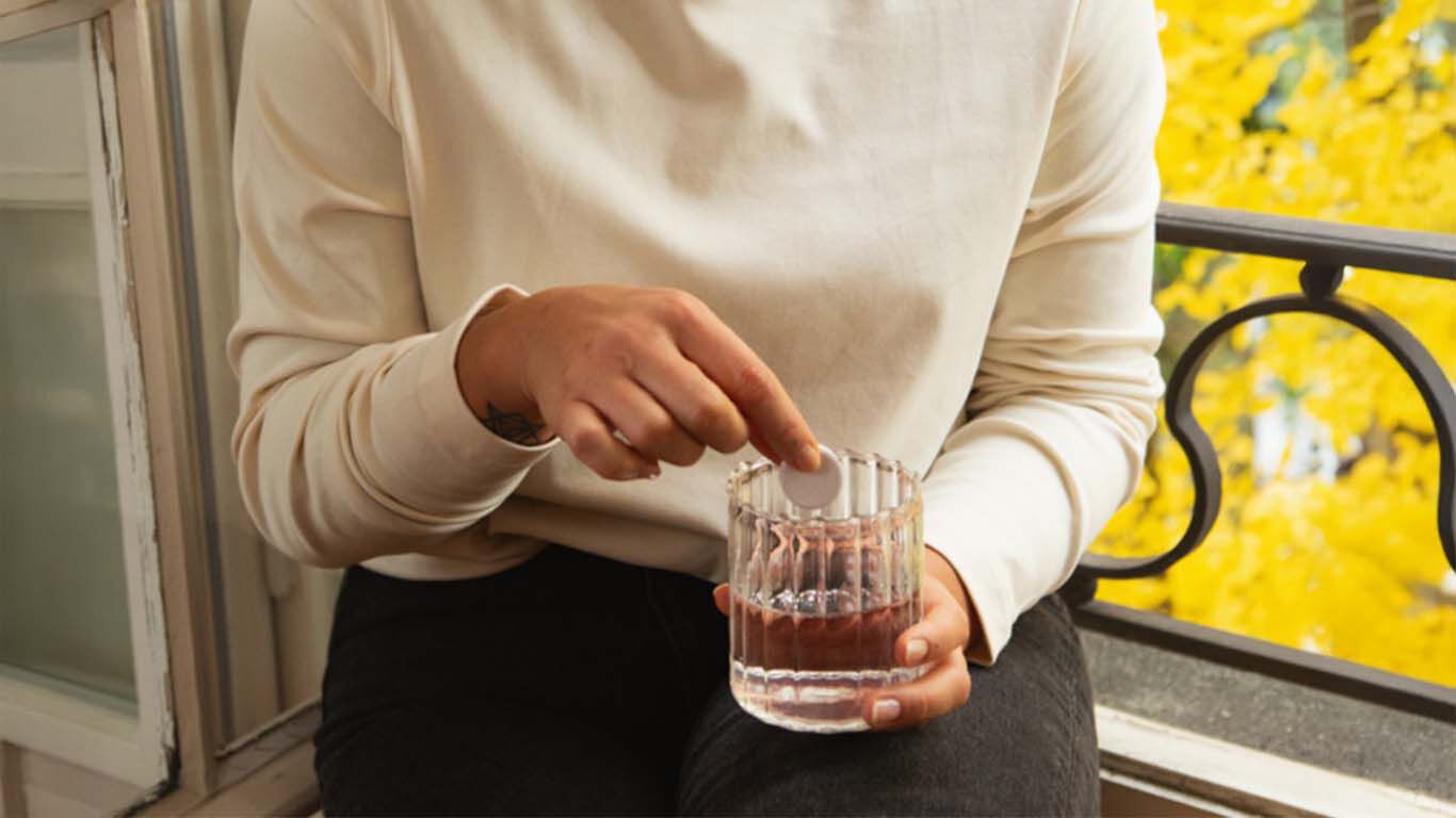 Femme tenant un verre d'eau et y dissolvant un comprim&eacute;, assise devant une fen&ecirc;tre avec un fond jaune automnal.