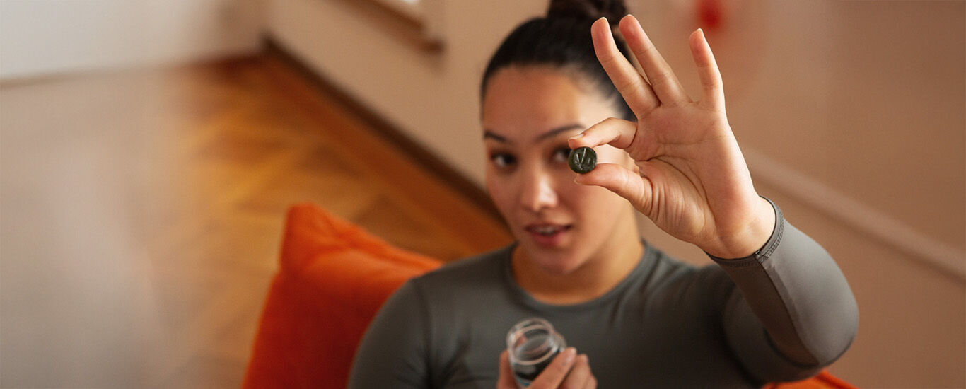 Une femme l&egrave;ve une main avec un gummie Kneipp vers la cam&eacute;ra et tient dans l'autre un emballage de gummie Kneipp, assise sur un canap&eacute; orange.