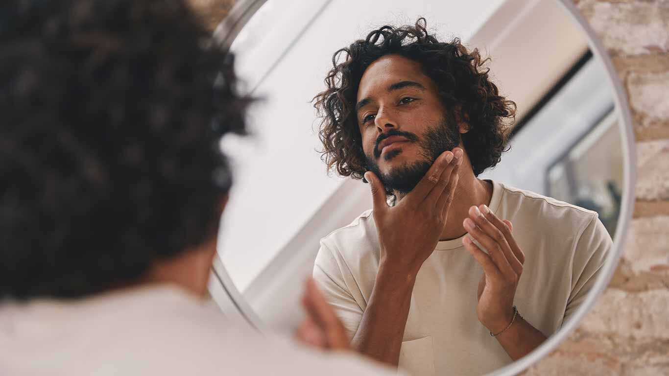 Un homme aux boucles sombres se tient devant le miroir et observe son visage. Une main touche sa barbe. 