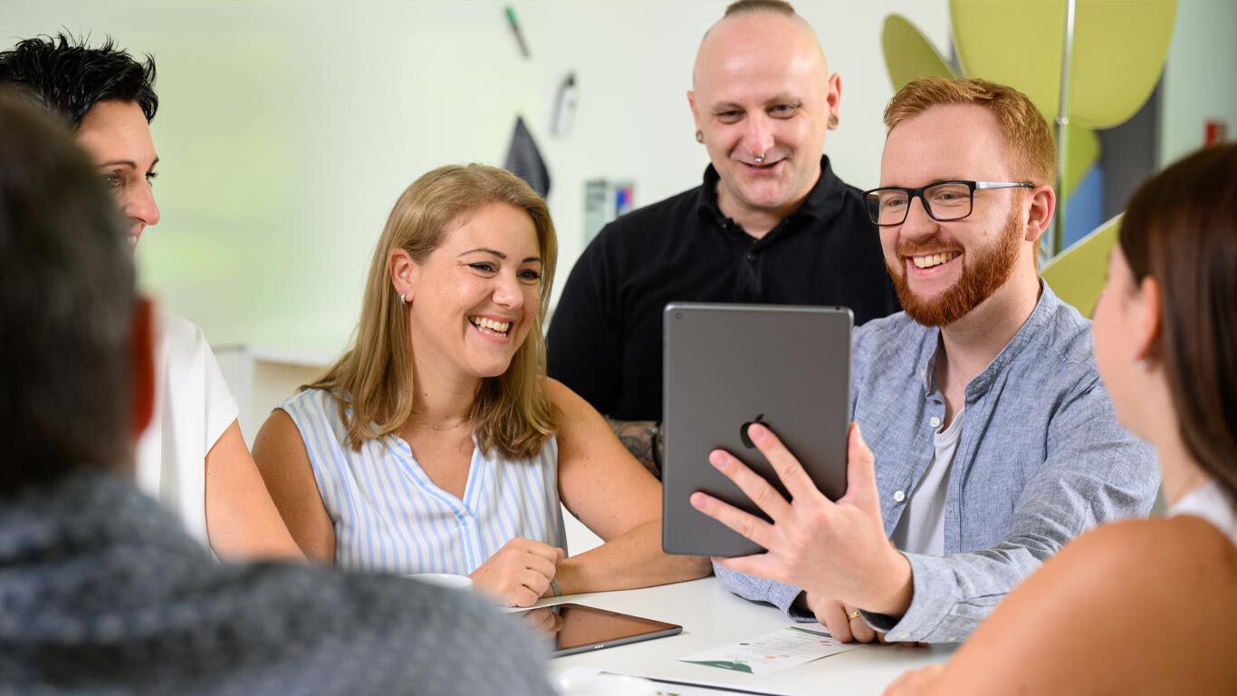 plusieurs personnes sont assises &agrave; une table, l'une d'entre elles tient une tablette en l'air, ambiance de travail, bonne humeur