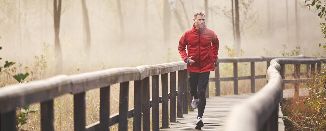Une personne v&ecirc;tue d'une veste de course rouge fait son jogging sur une passerelle en bois au milieu d'une for&ecirc;t brumeuse.