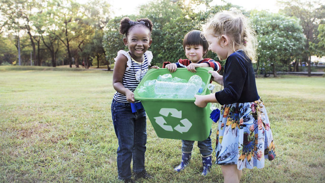 Trois enfants dans un pré avec une poubelle verte remplie de bouteilles en plastique