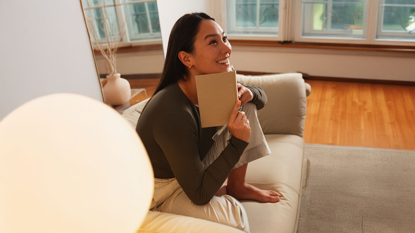 brune est assise sur le canap&eacute; et tient un livre &agrave; la main en riant.
