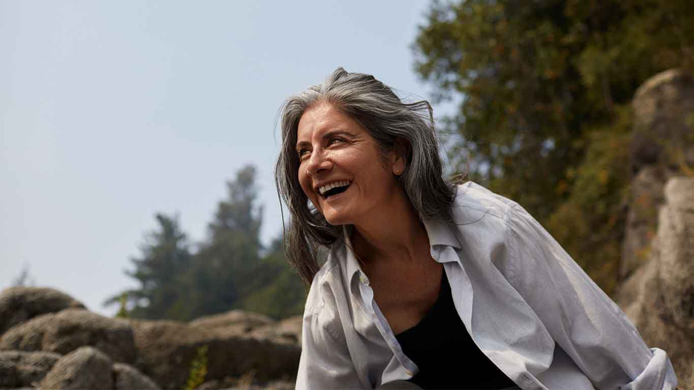 Une femme aux cheveux gris et au chemisier blanc grimpe joyeusement du haut d'un rocher. 