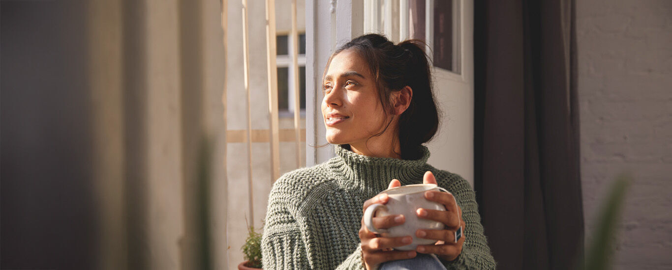 Femme assise &agrave; la fen&ecirc;tre avec un pull-over vert en tricot et une tasse &agrave; la main, regardant dehors.