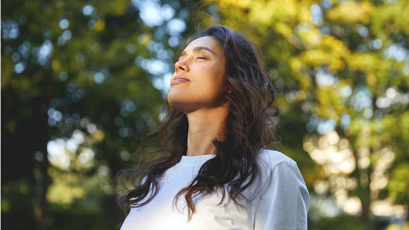 Une femme en T-shirt blanc se tient dans un parc et, les yeux fermés, profite du soleil sur son visage. 