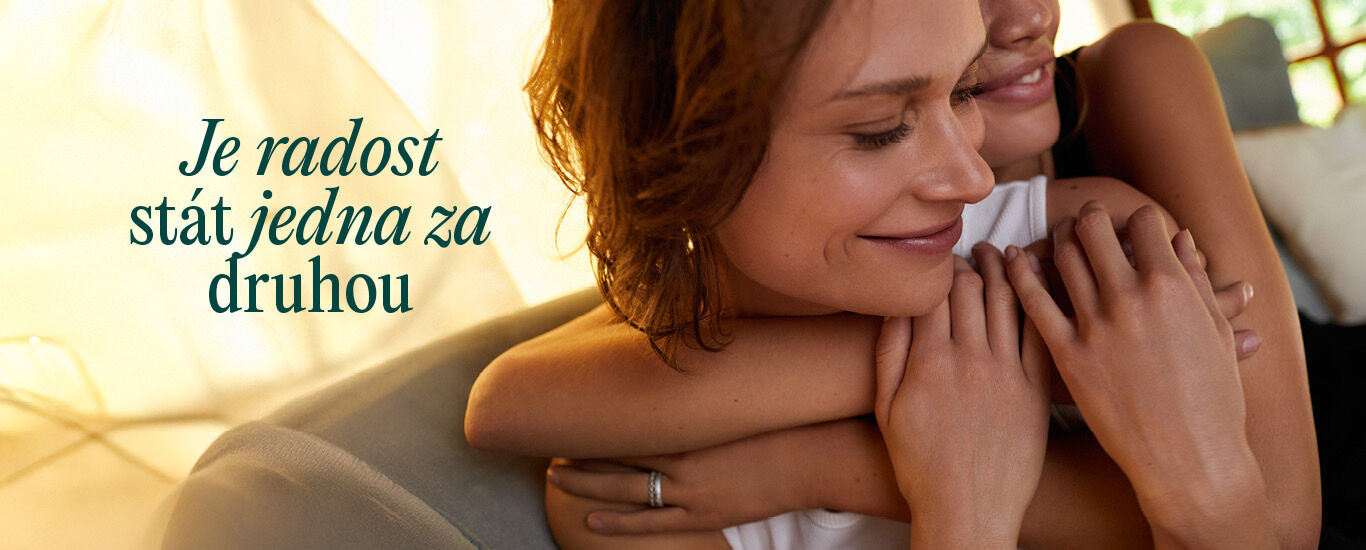 Žena objímá druhou ženu, obě se usmívají.