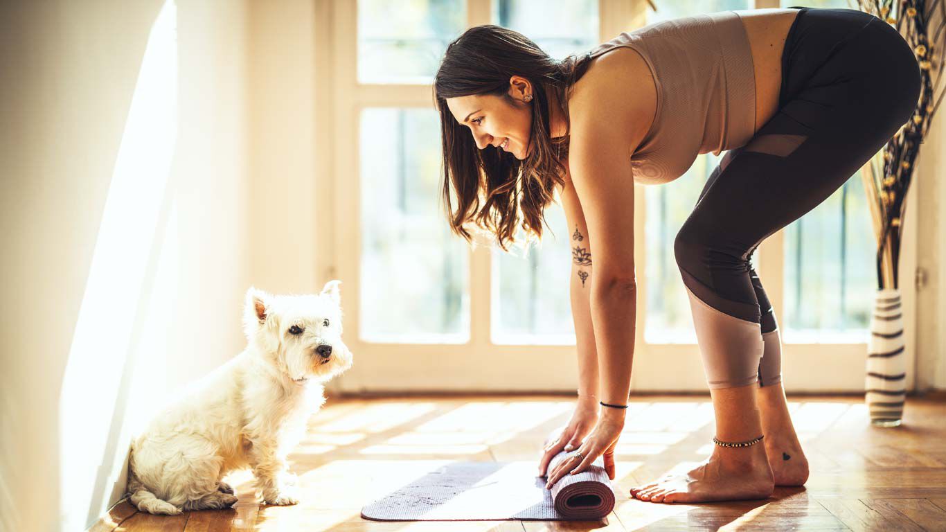 Mujer enrollando la esterilla de yoga, un perro blanco al lado.