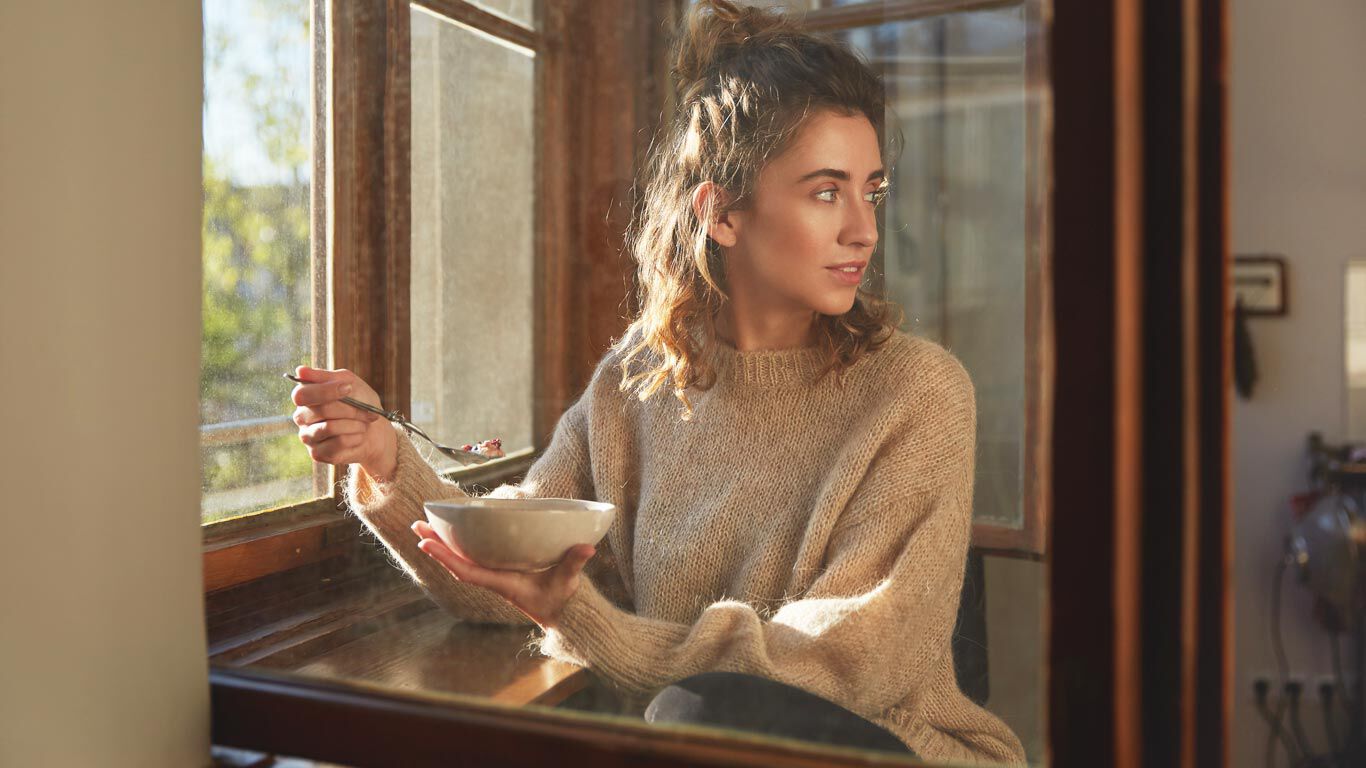 Mujer sentada junto a la ventana comiendo cereales.