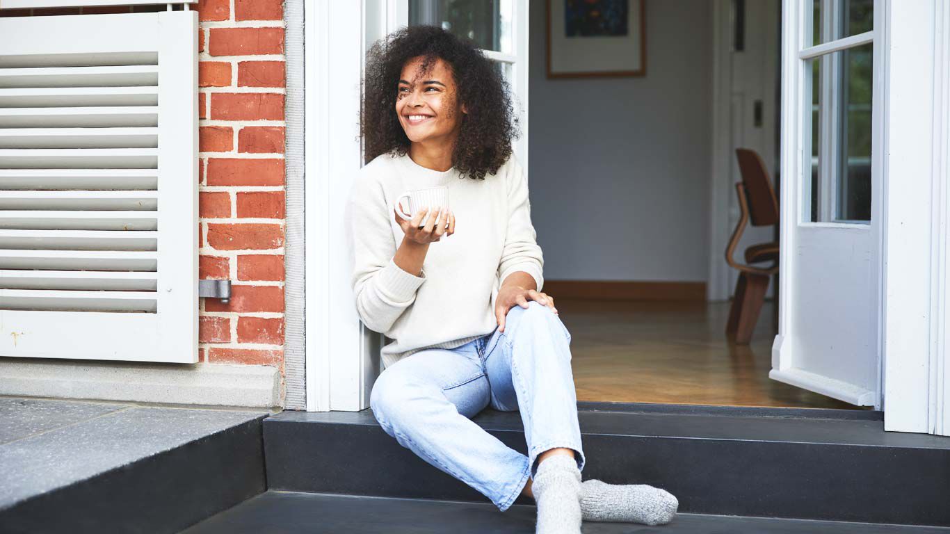 Femme heureuse assise à la porte de la véranda, une tasse à la main.