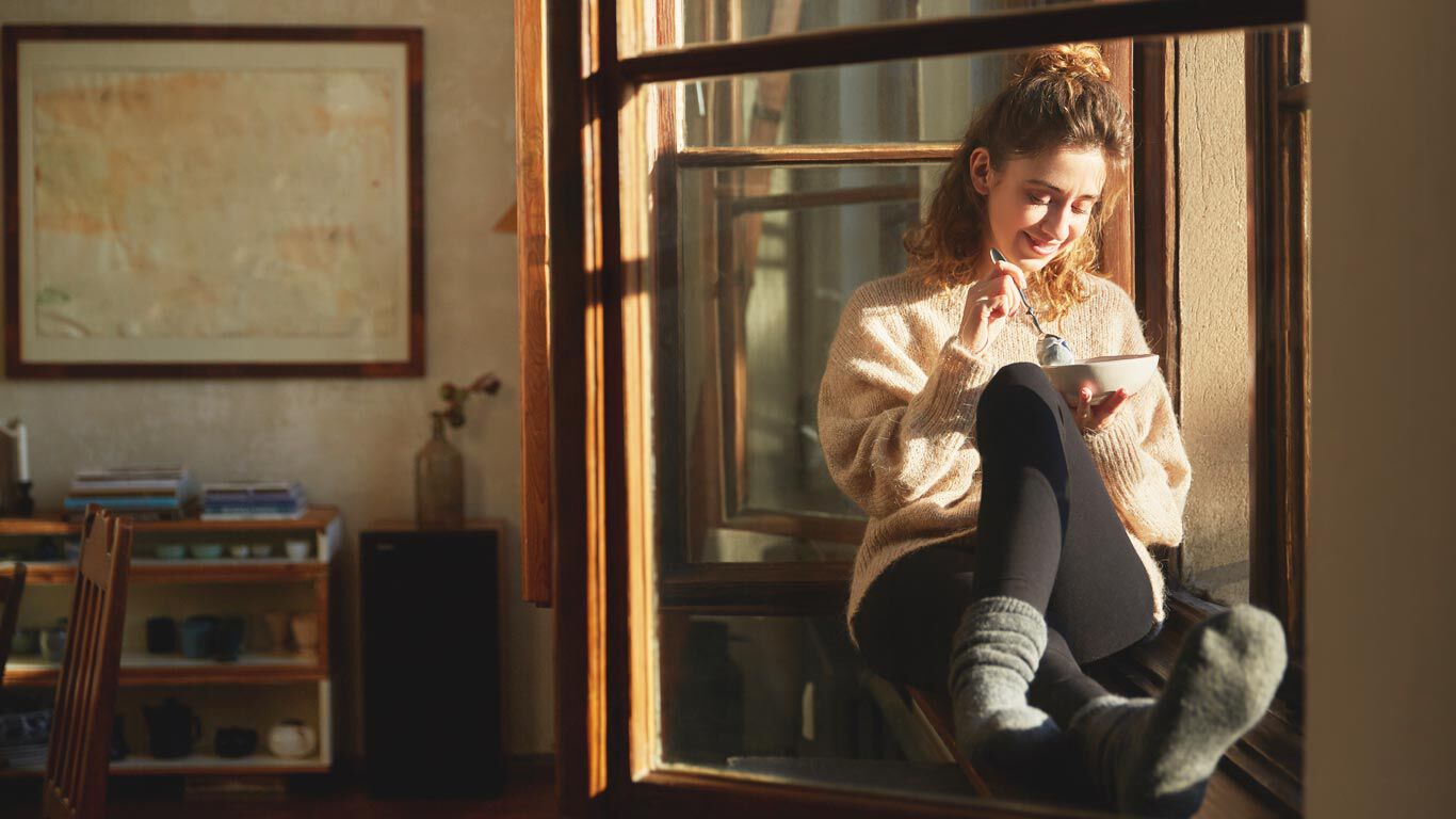 Une femme est assise sur le rebord de la fenêtre dans des vêtements confortables et mange des céréales.